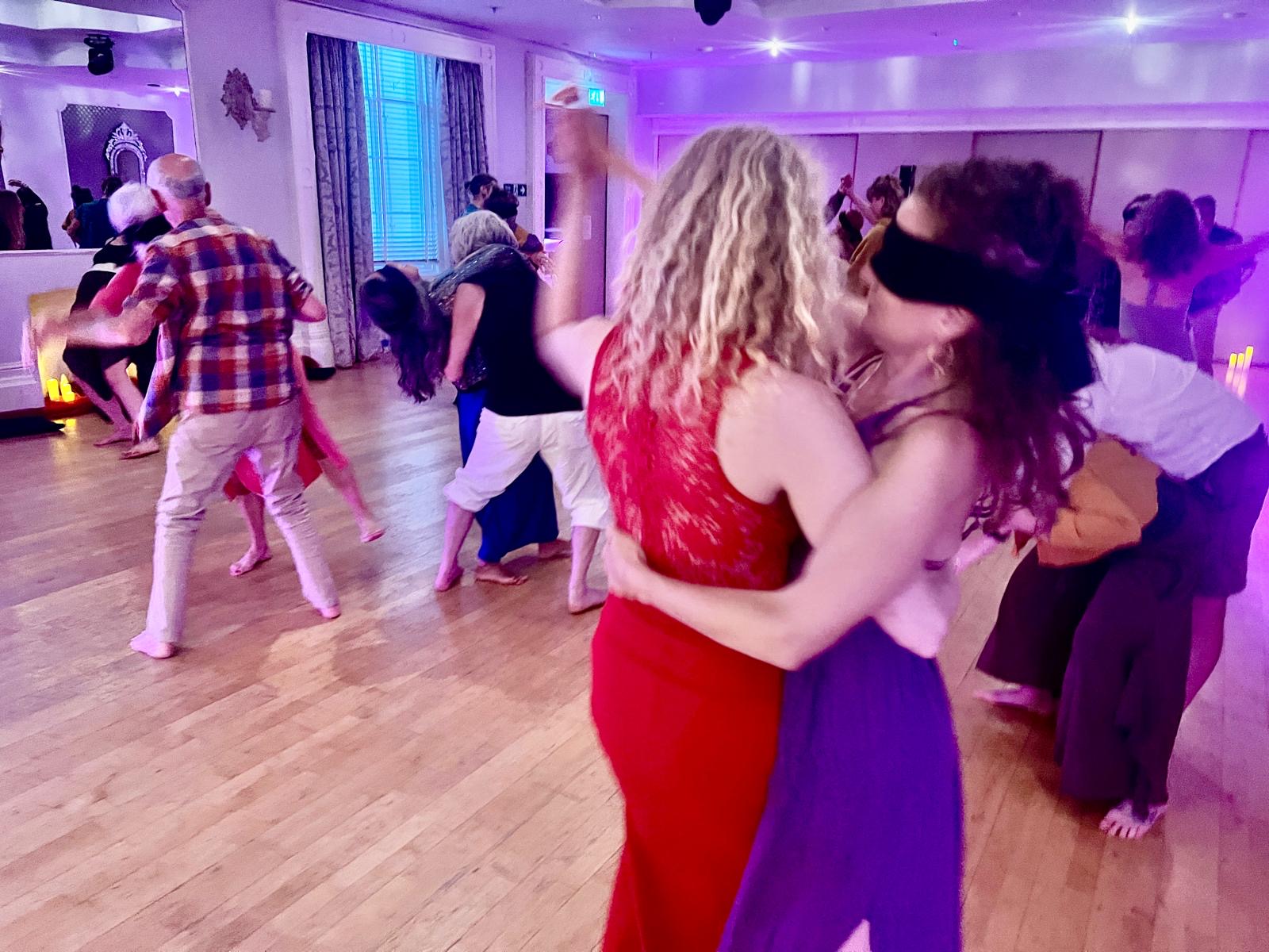 Two women dancing in a close embrace in the foreground, one blindfolded, while the group moves behind them
