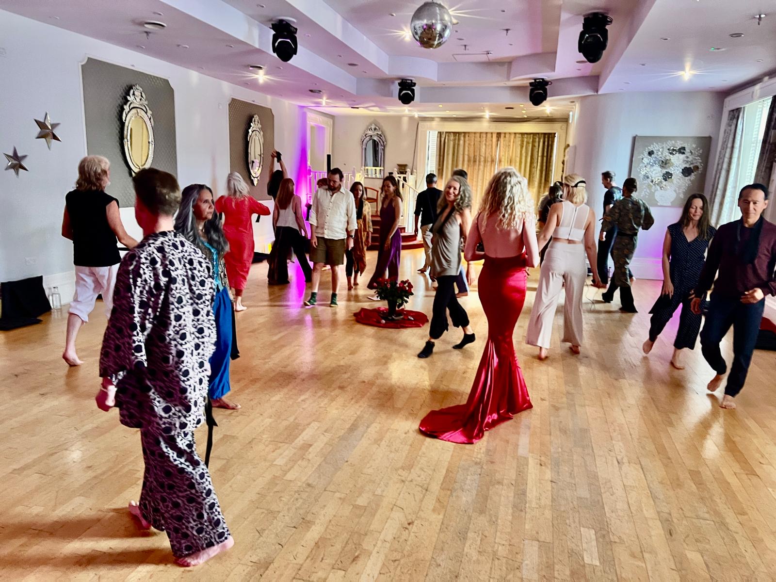 A wide view of the dance floor: participants walking and moving freely around a centrepiece of roses, a woman in a long red dress at its heart