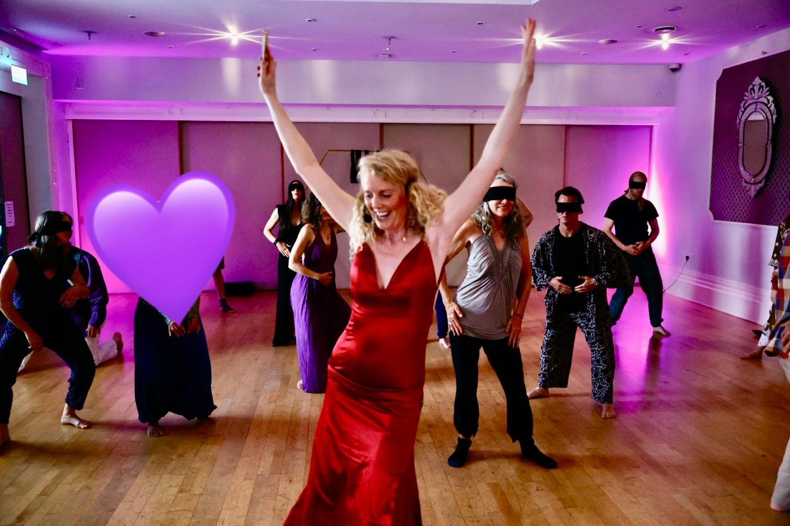 A dancer in a red dress with both arms raised in joy as the group moves around her